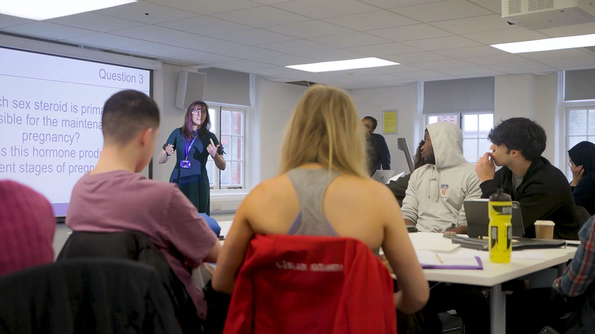 An example of larger group CBL teaching being led by a facilitator. A group of students are sat around a table in a large classroom observing a screen. The facilitator is stood next to the screen detailing various points relating to the case in question which is being projected onto the wall.