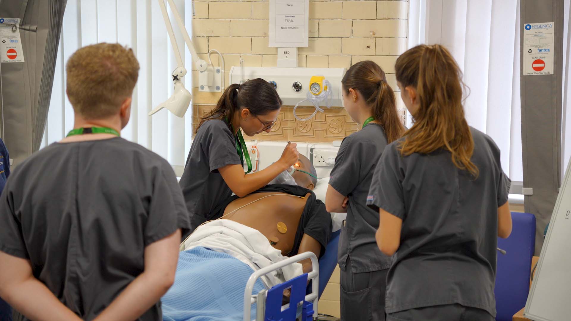 A wide shot of medical students crowded around a patient bed in a simulated ward environment