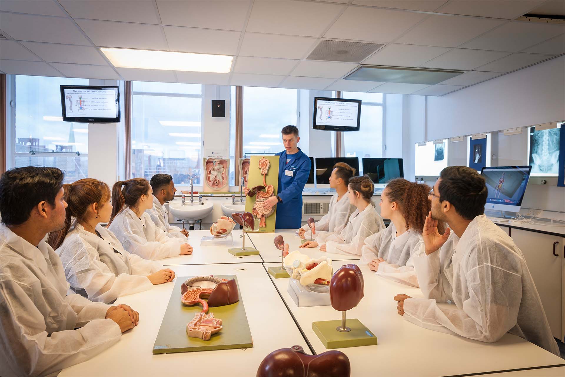 A shot of medical students studying in an anatomy class - they are all wearing white lab coats. In the middle of them stands a clinical academic with a model of the human body.