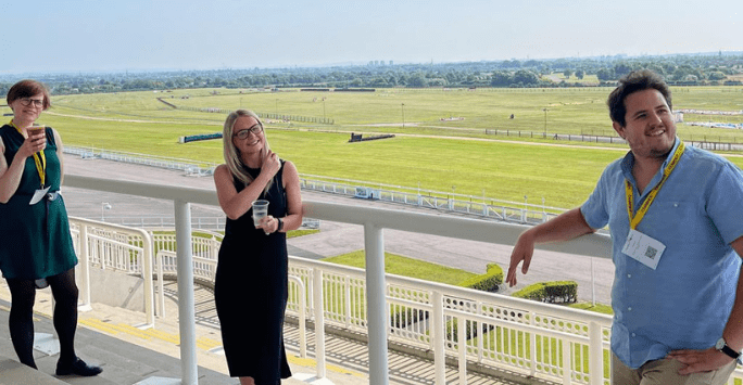 3 people stand on a viewing platform with a green racetrack and hills in the background