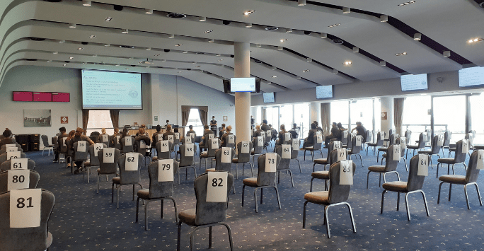 students sit on numbered chairs in a room with large windows watching a presentation