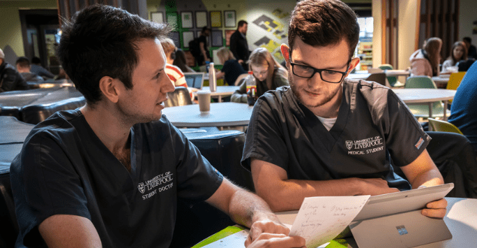 two students in grey scubs study with a tablet and written notes