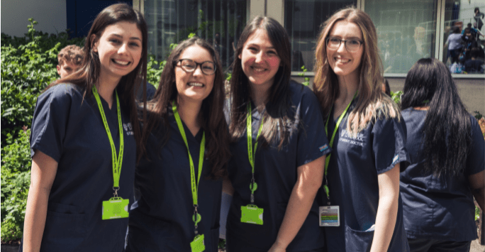 Four women in grey scrubs smile to the camera