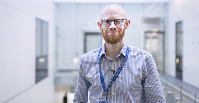 man wearing glasses, a shirt and lanyard smiles in a bright indoor space