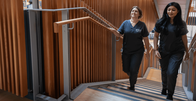 two student doctors in grey scrubs walk up a modern flight of stairs