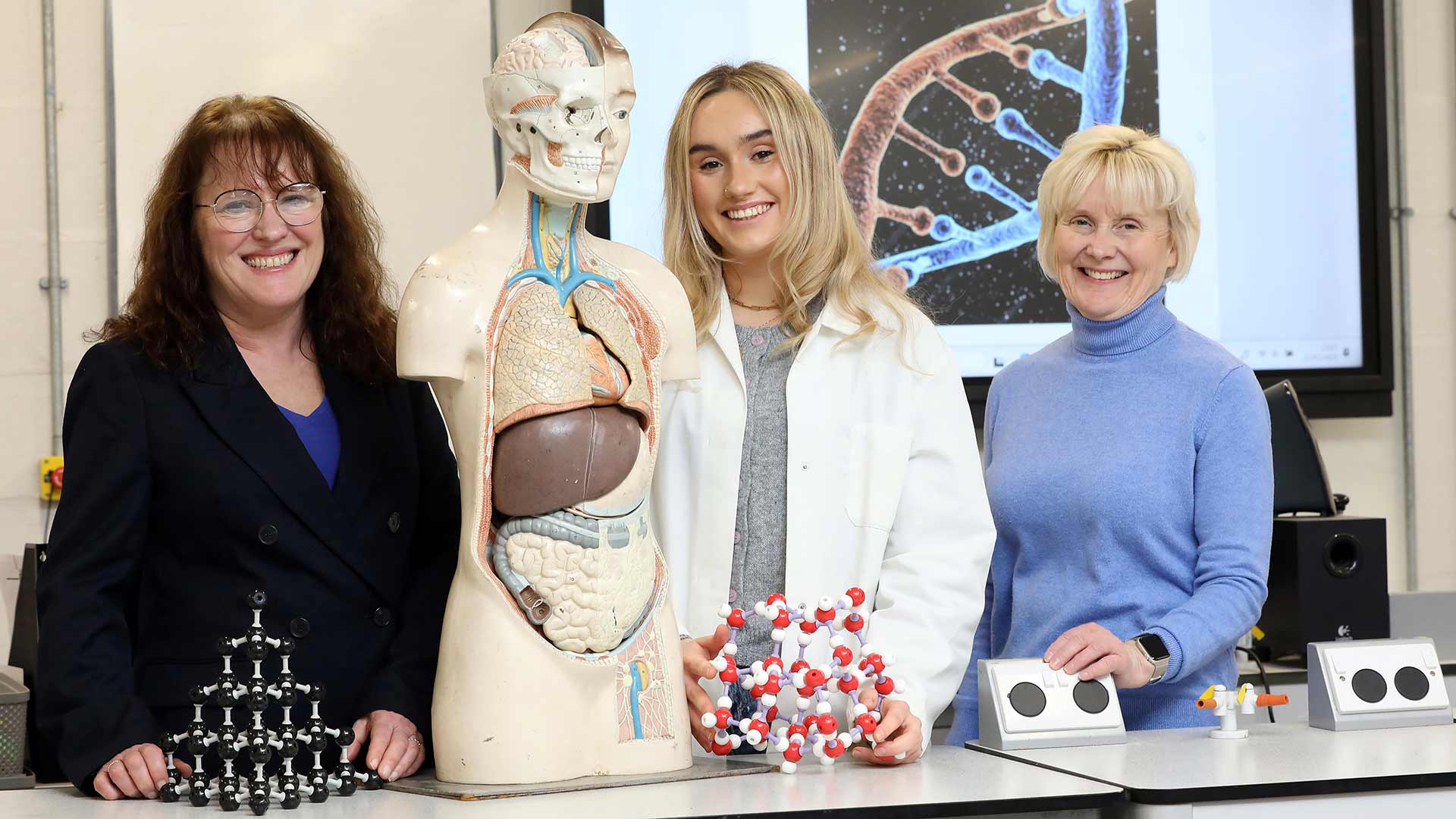 student in white lab coat poses alongside anatomy equipment with two educators