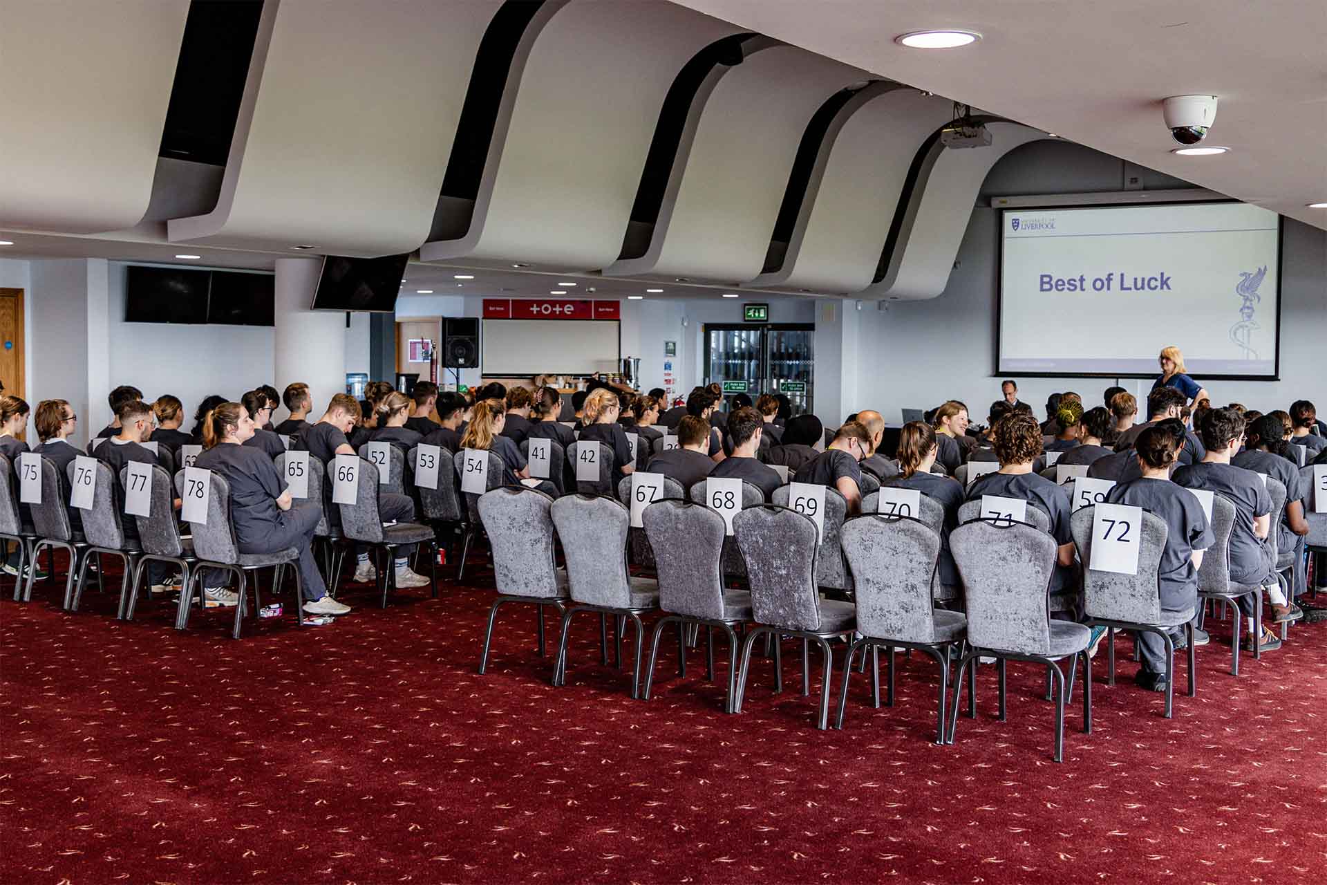 An exam hall full of students sat in chairs facing a projector. The text on it reads 