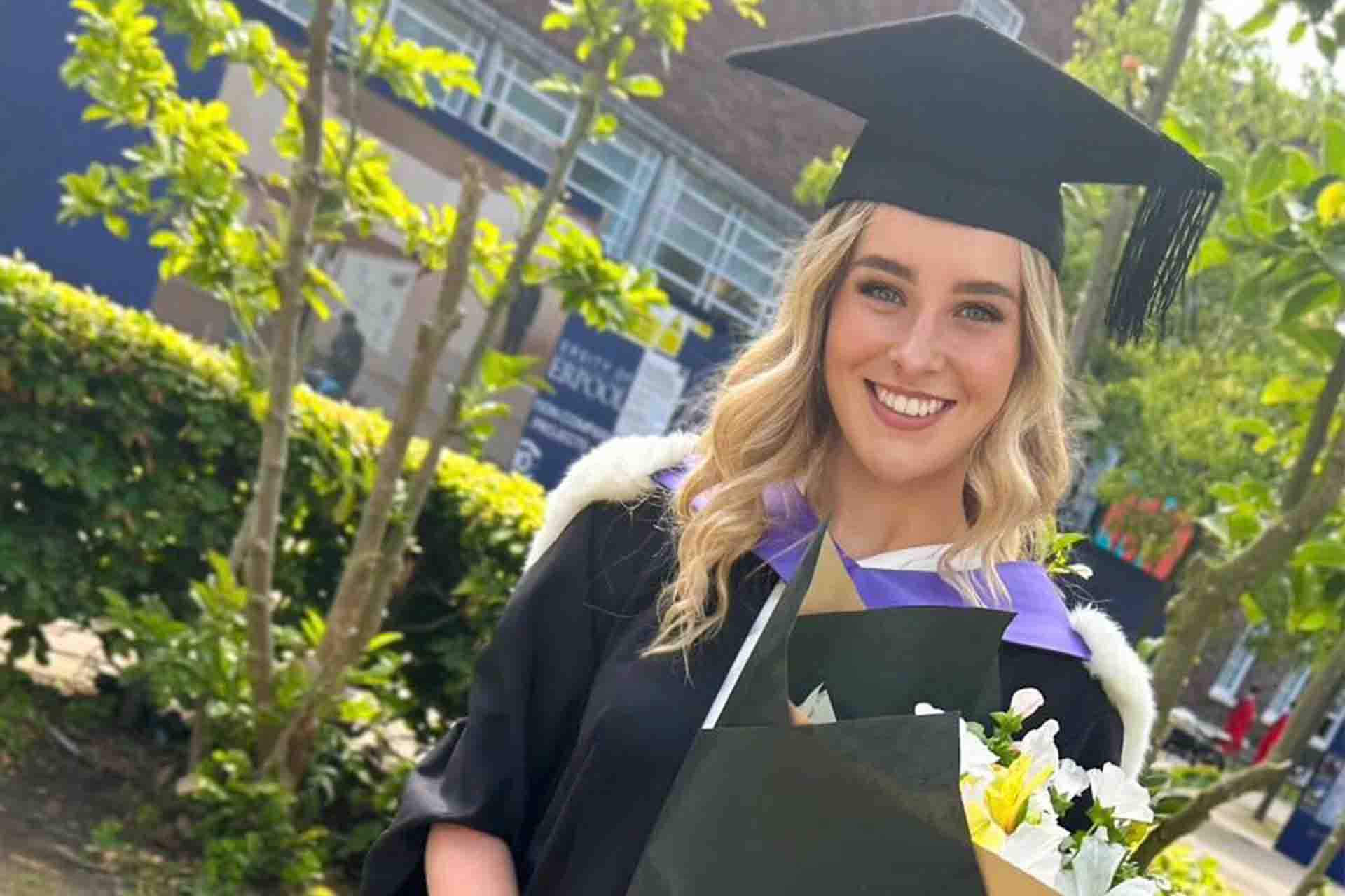 a graduate in cap and gown with greenery behind