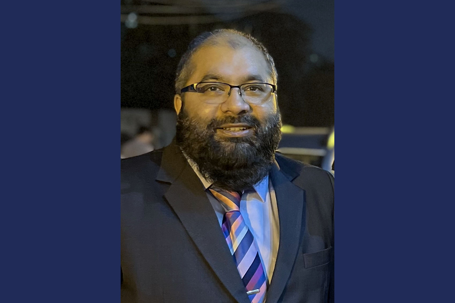 A profile head shot of a man on a blue background. He is wearing glasses, has a full black beard, and is in a grey suit with a blue tie.