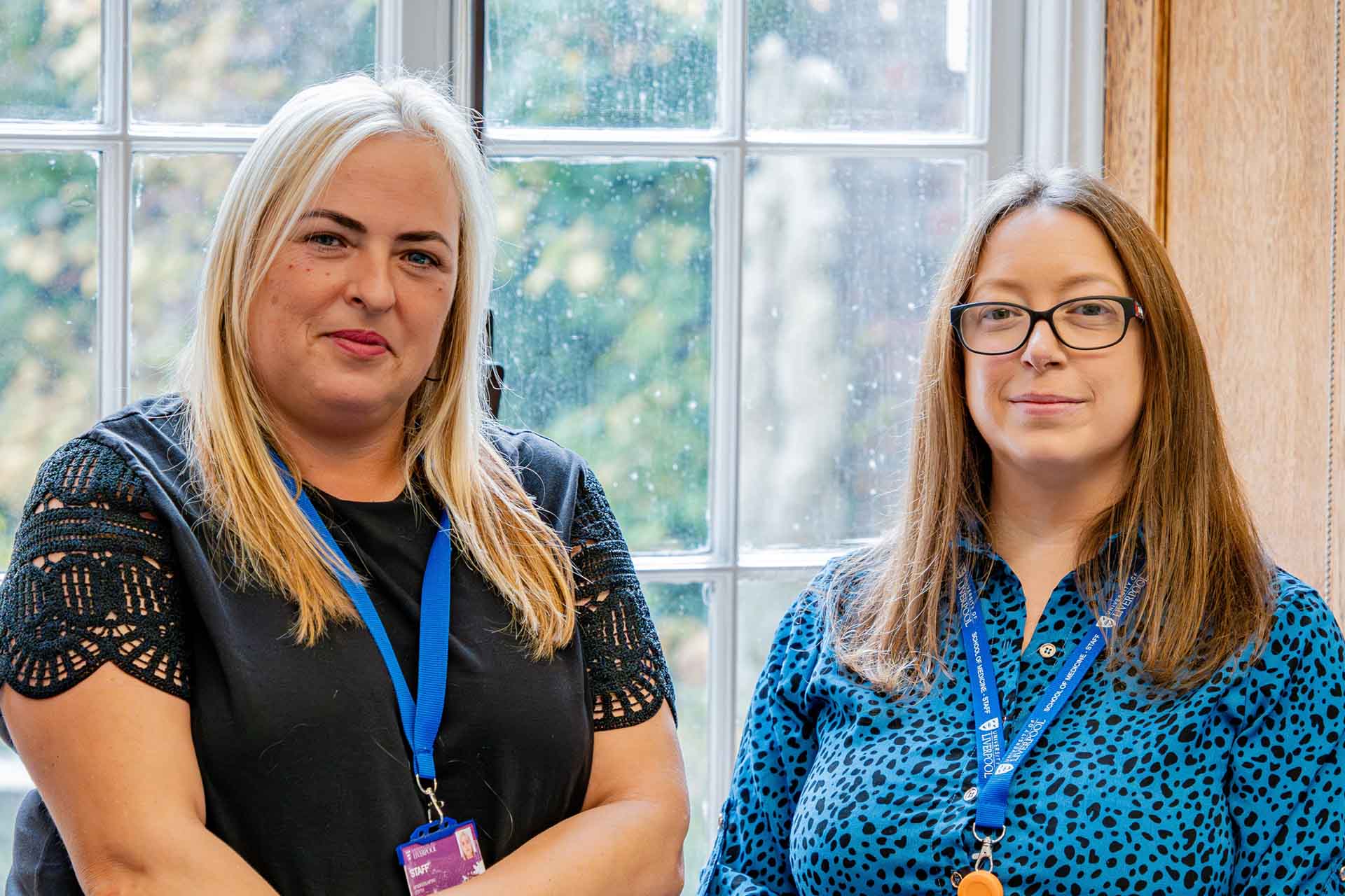 An image of two women stood in front of a wooden window frame. The woman on the left is slightly taller, with blonde hair & dressed in a black top. The woman on the right is wearing glasses, has brown hair & in a blue top.