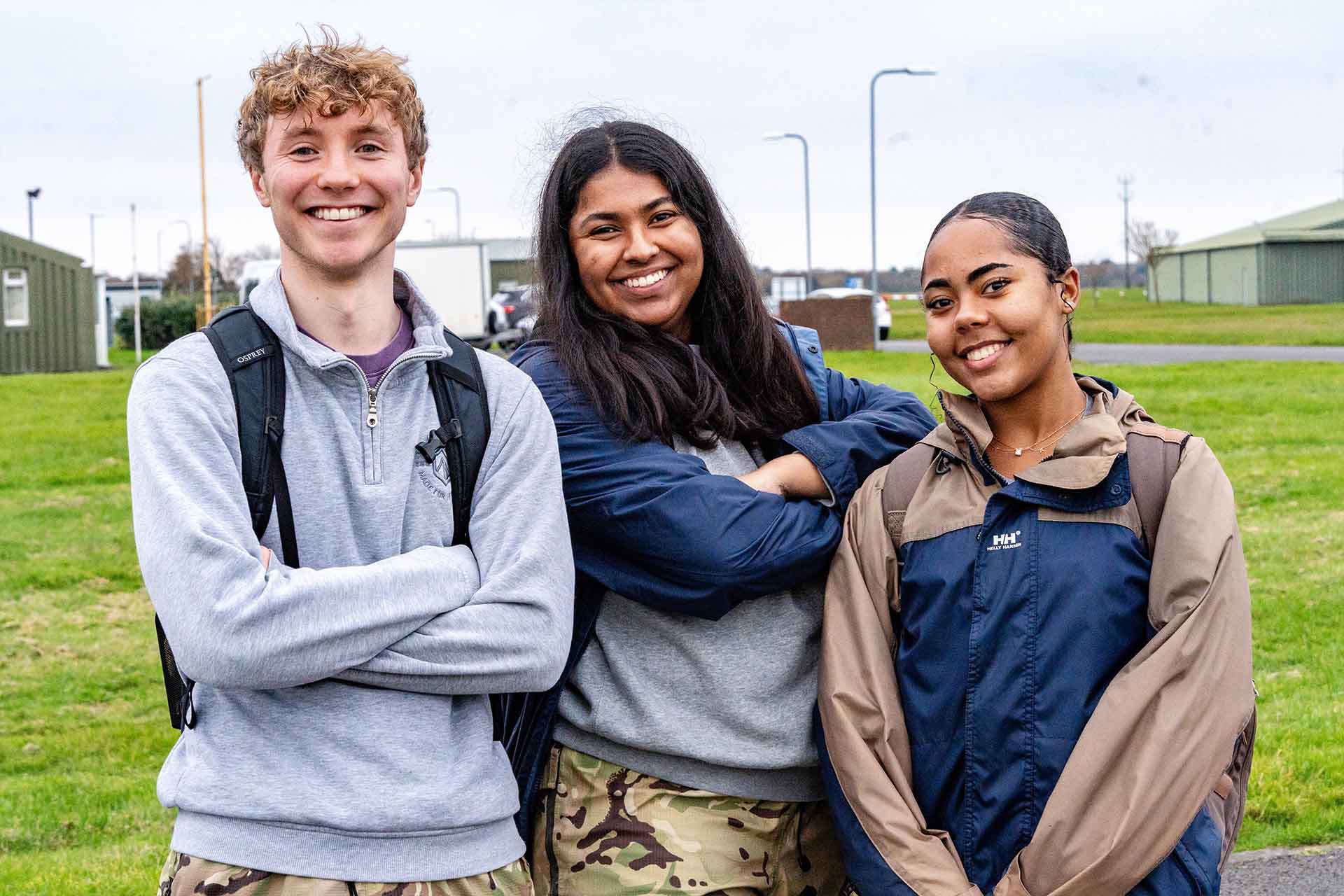 Three students facing camera and smiling. The leftmost is male and the other two students are female. Behind them is an RAF base.