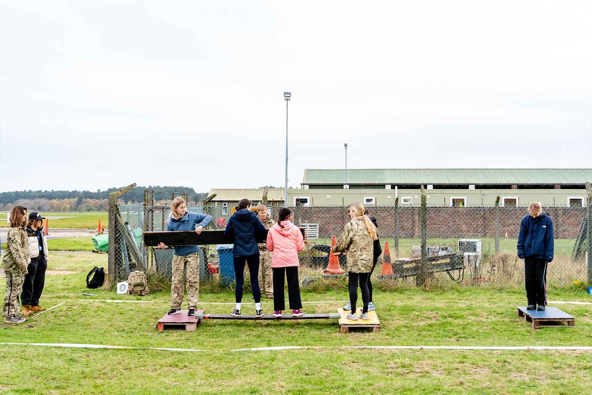 A group of students stood in an open field on two raised pallets. They are in separate groups on each pallet and passing a wooden board between them in order to convey the group to the other side.