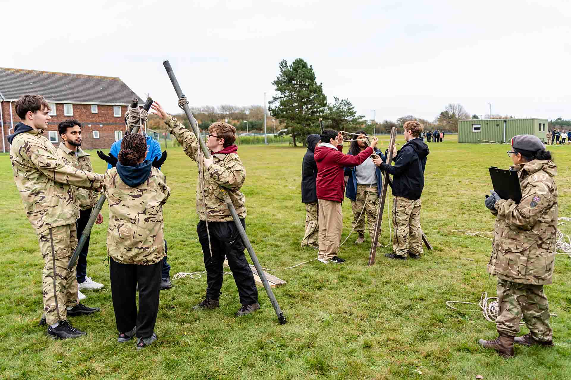 Two groups of students working at a problem outside. One group is tiding together three long metal poles with a piece of rope in a triangle, the other is constructing a wooden triangular base to attach to this structure.