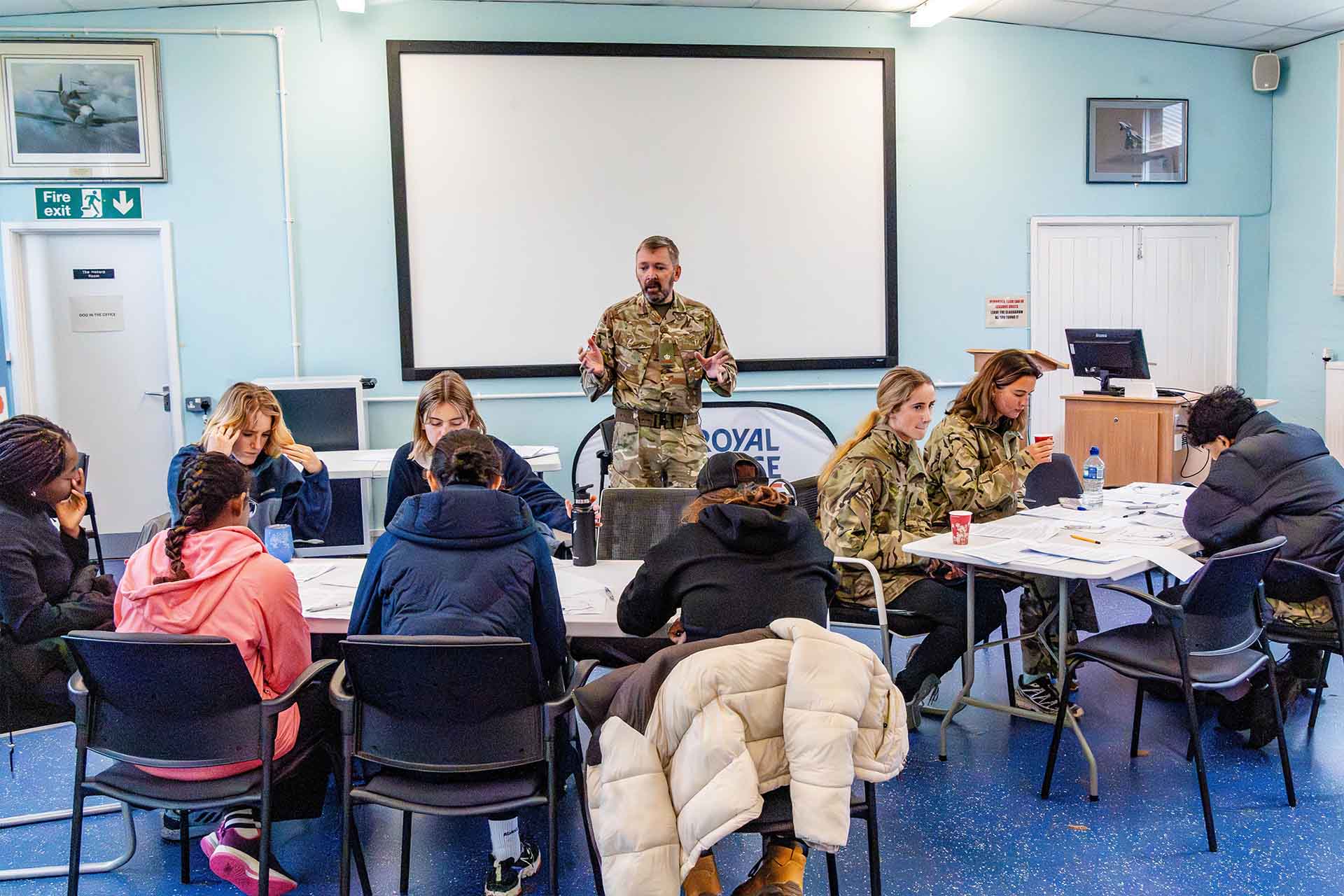 A group of students sat in a classroom at tables. In front of them is an RAF instructor leading a planning session.