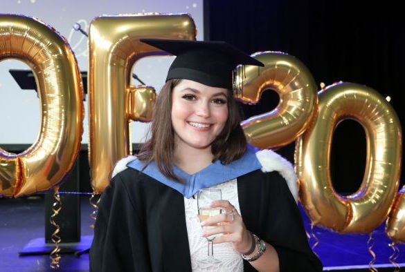 Student Beth Whitham wears a blue and black gown and holds a drink while smiling