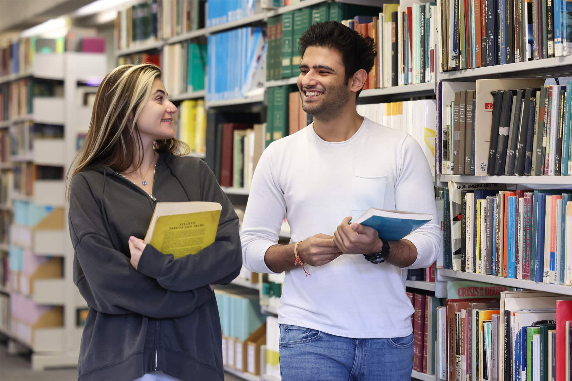 Two students carrying books, walk past a book shelf filled with books.