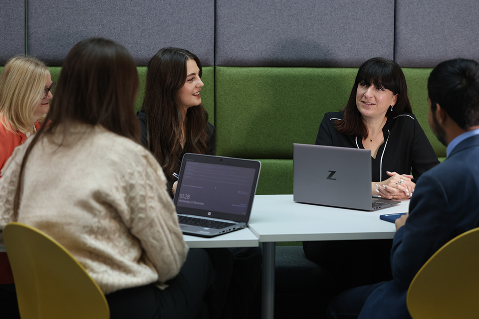 Group of staff sitting at a table with laptops, talking to each other