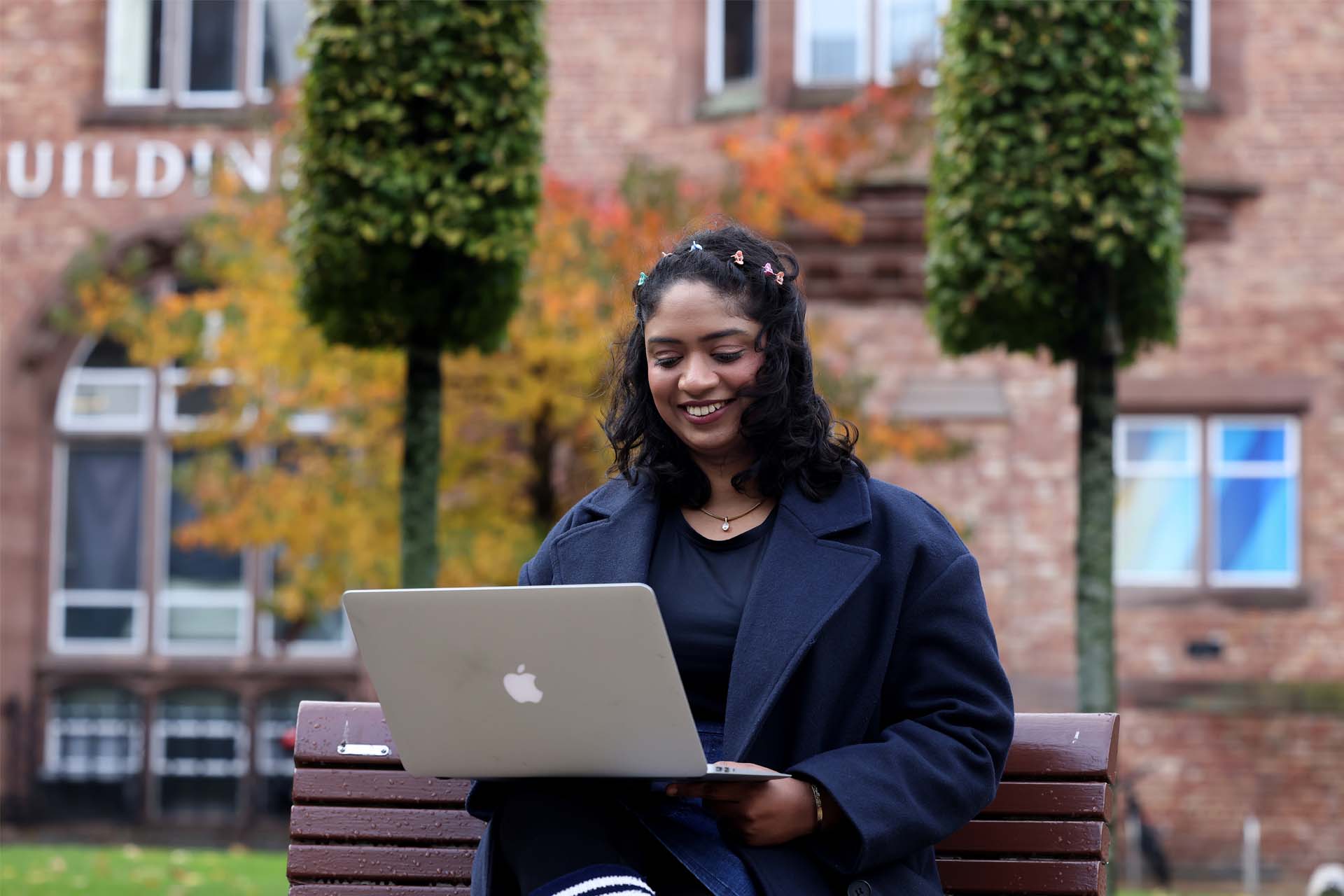 A student sitting outside on a bench on campus using a laptop.