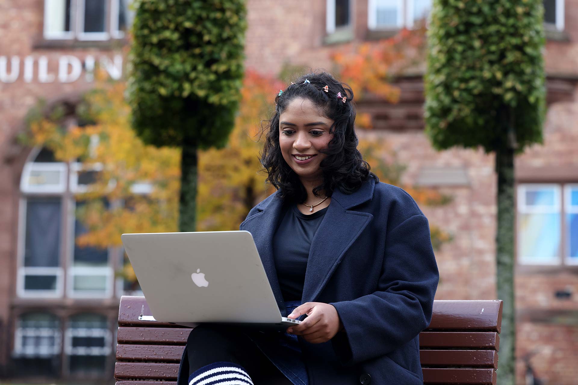 A student works on a Macbook while sitting on a bench in a courtyard on campus.