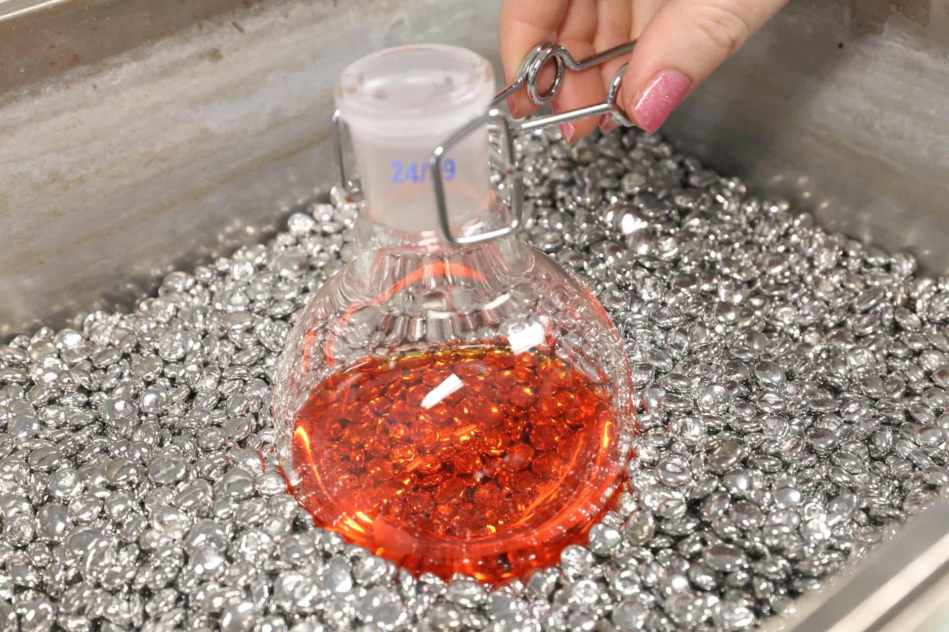 A beaker is being held in a tank of silver rocks by hands with pink sparkly nail polish.