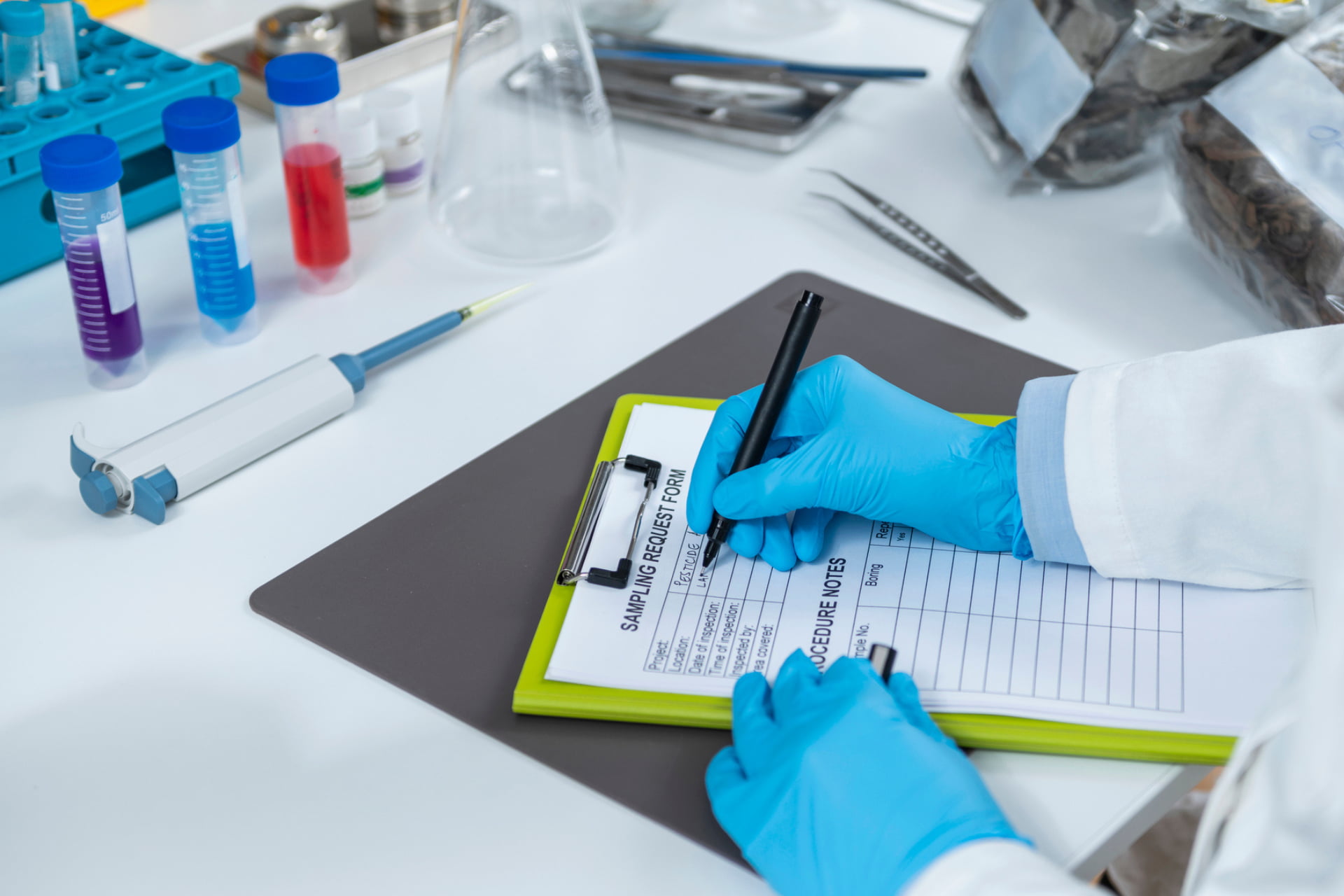 On a lab table full of lab equipment, a hand wearing blue plastic gloves writes on a green clipboard.