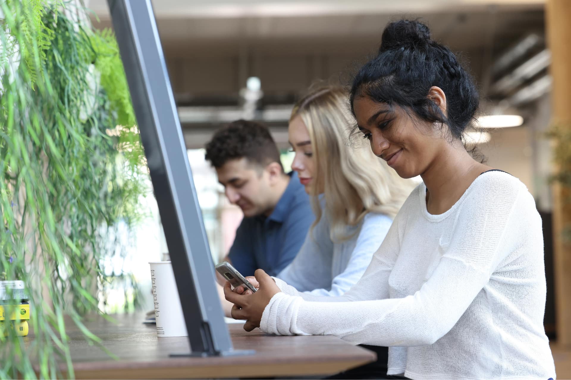 A student with black hair sitting at a table with other students smiling and looking at her phone.