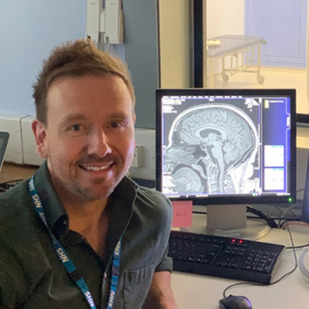 A picture of Simon Keller with a computer monitor displaying a brain scan in the background