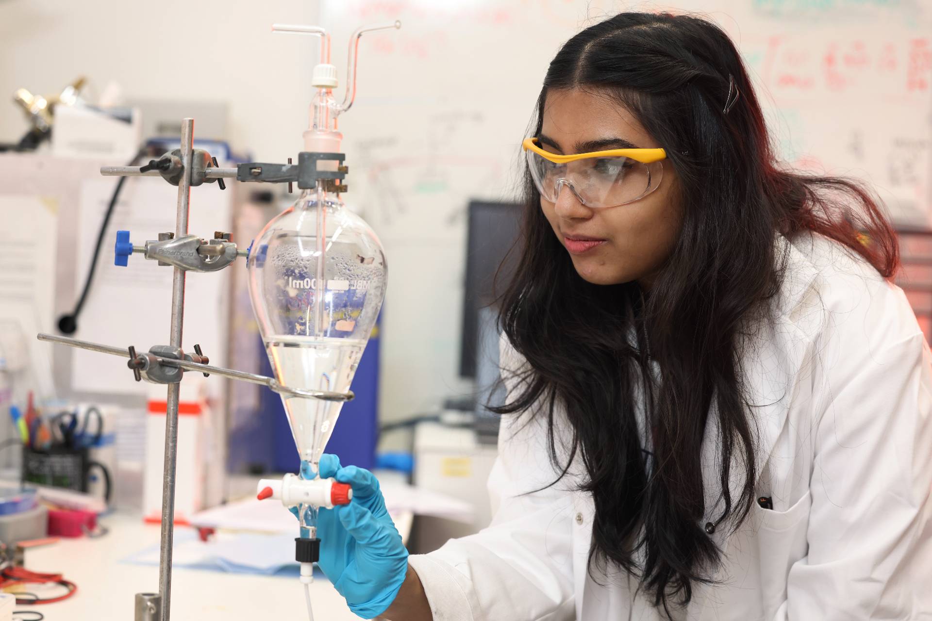 A female student wearing safety goggles and a lab coat works in a lab.